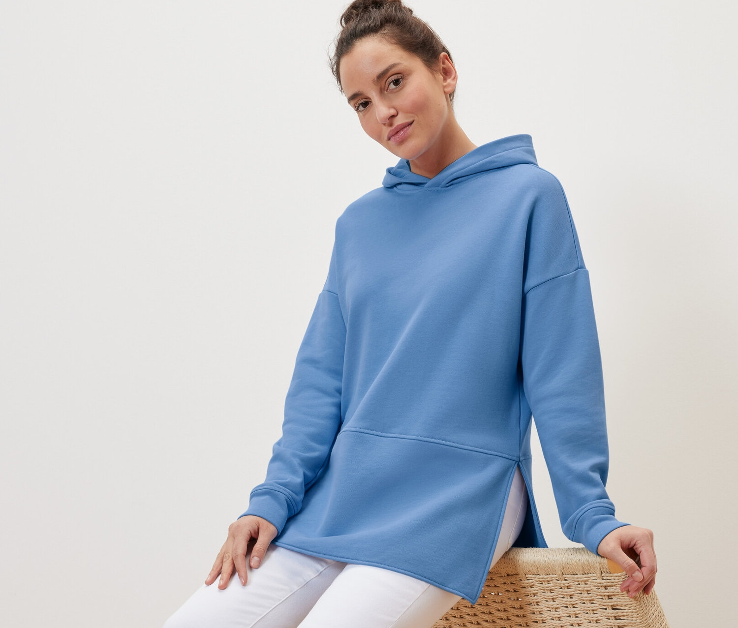 Žena sedí na prútenom kresle a má na sebe svetlomodrú dlhú mikinu s kapucňou.