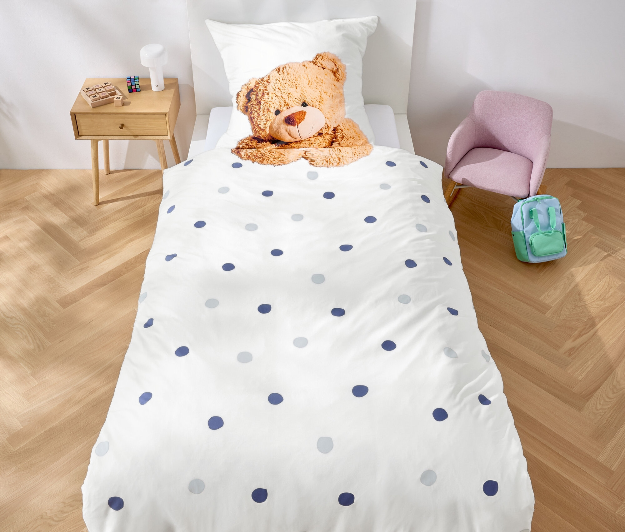 V izbe stojí posteľ s bielymi obliečkami s modrými bodkami a plyšovým medvedíkom. Nad posteľou visí girlanda z fialových trojuholníkov.