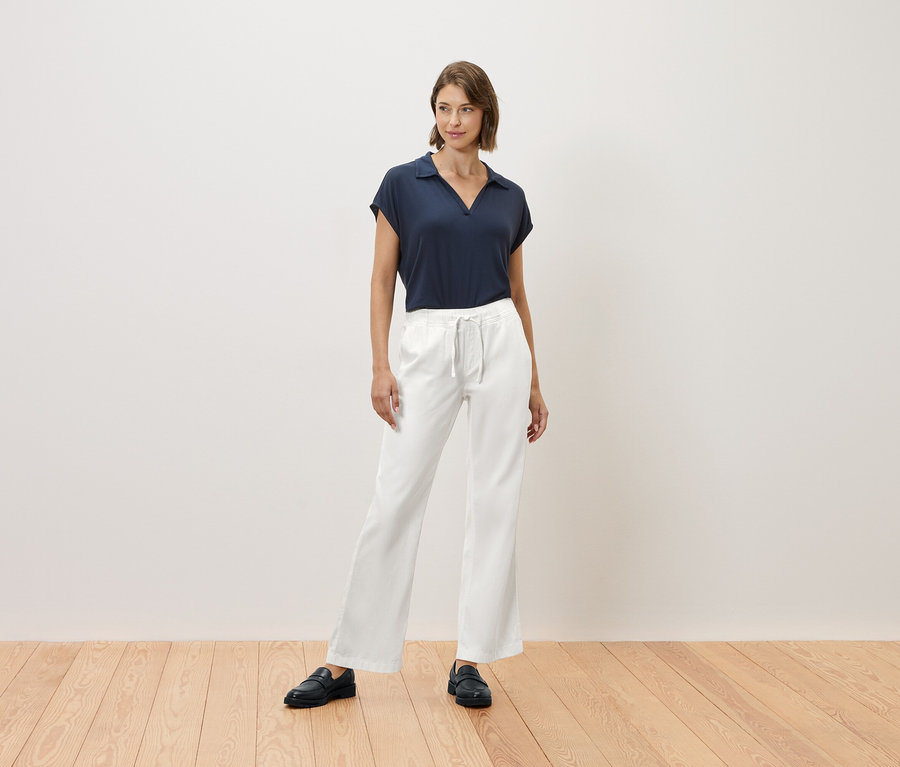 Žena stojí na drevenej podlahe a má na sebe tmavomodré tričko a biele nohavice.