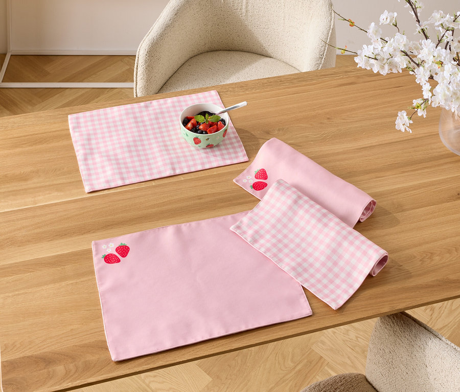 Na drevenom stole sú ružové prestierania, jedno s jahodovým vzorom, miska s bobuľami a váza s kvitnúcimi vetvičkami.