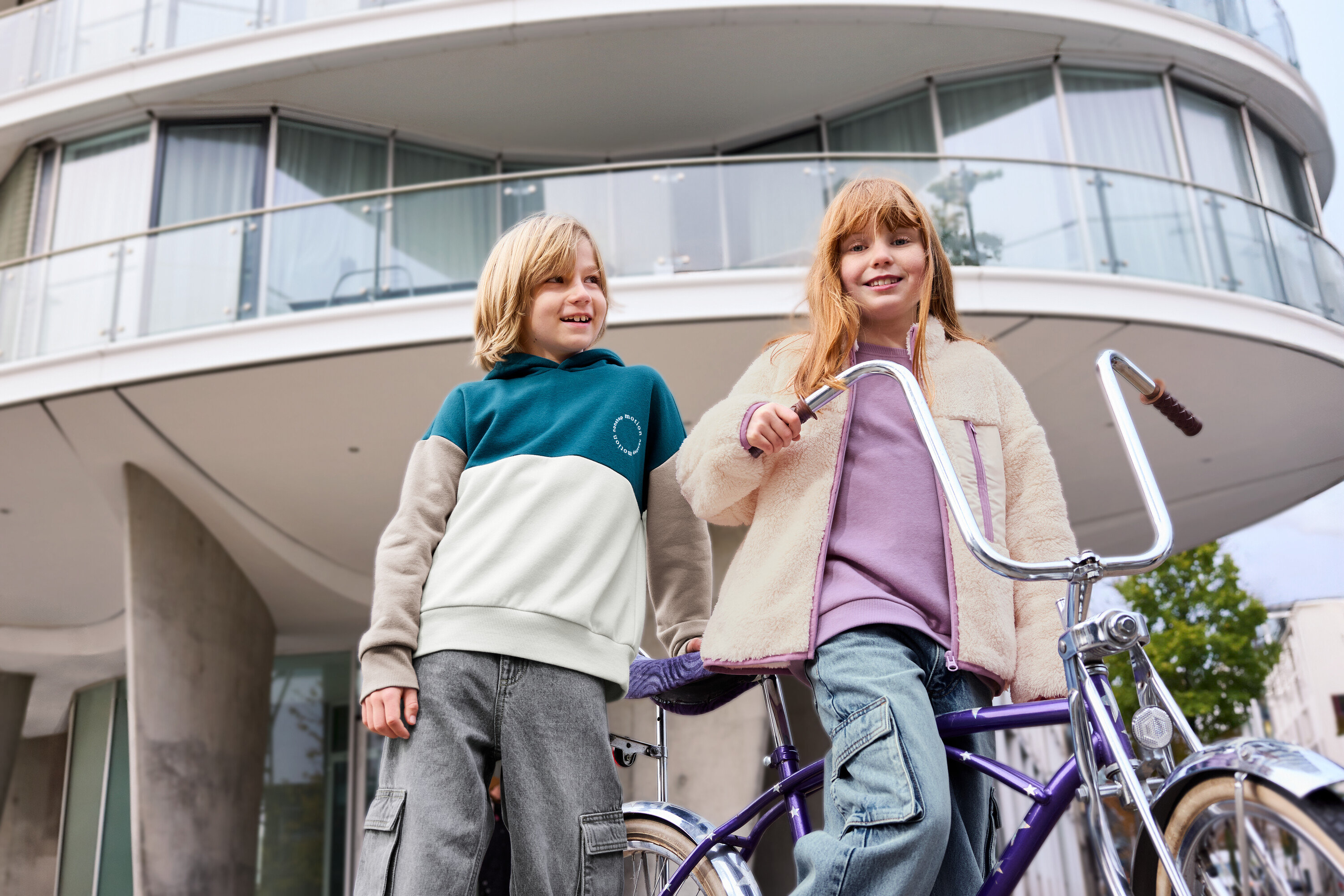 Dve deti stoja pred budovou, jedno dieťa má na sebe levanduľovú detskú mikinu s počesanou vnútornou stranou.