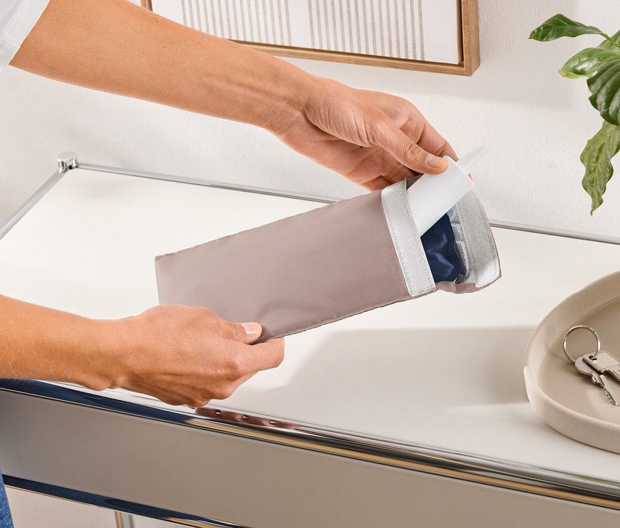 Osoba otvára podlhovastý, béžový ochranný obal, ktorý obsahuje modrý produkt.