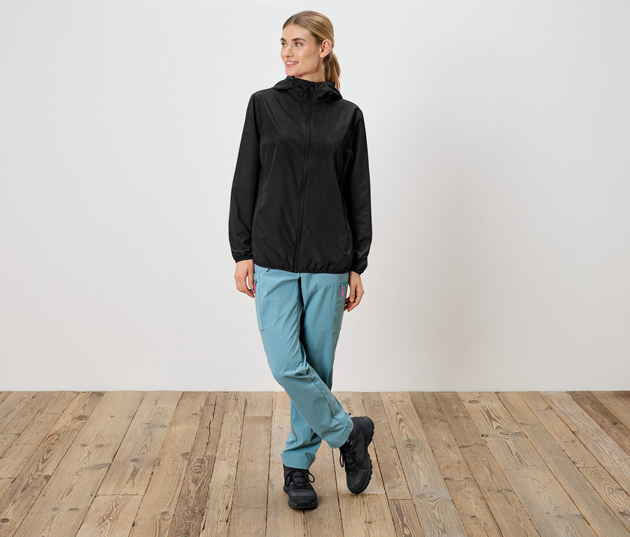 Žena v čiernej nepremokavej bunde s kapucňou, svetlomodrých nohaviciach a čiernych topánkach stojí pred bielou stenou.