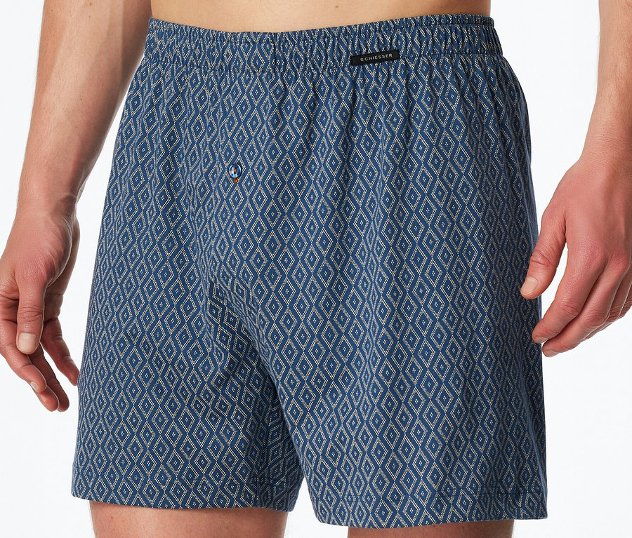 Muž má na sebe modré džersejové boxerky SCHIESSER s kosoštvorcovým vzorom a zapínaním na gombík.
