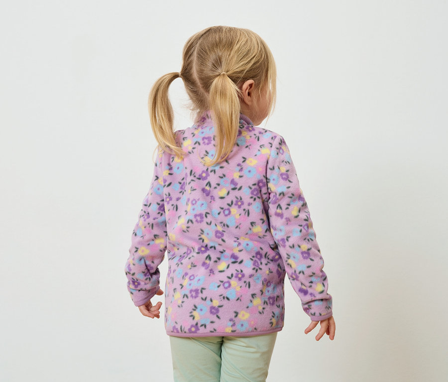 Pohľad zozadu na malé dievča s blond vrkočmi, oblečené v lila fleecovej bunde s kvetinovou potlačou a zelených nohaviciach.