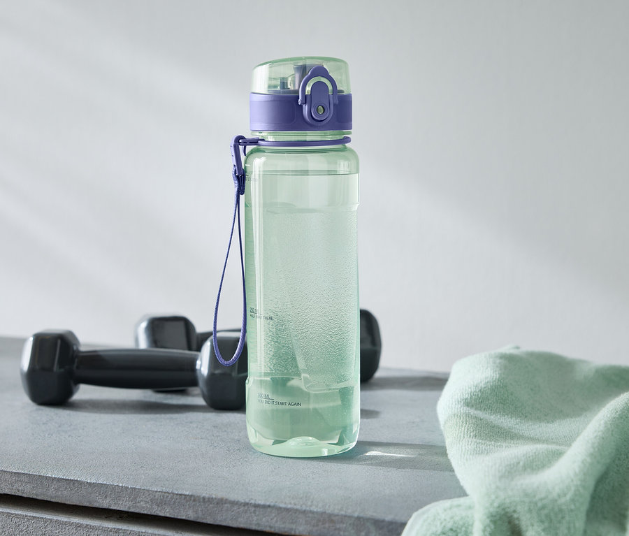 Zelená fľaša na vodu s fialovým uzáverom stojí vedľa dvoch čiernych činiek a svetlozeleného uteráka na sivom povrchu.