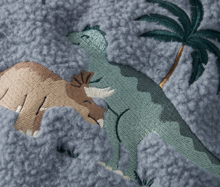 Detail šedej látky s vyšitými dinosaurami a palmou.