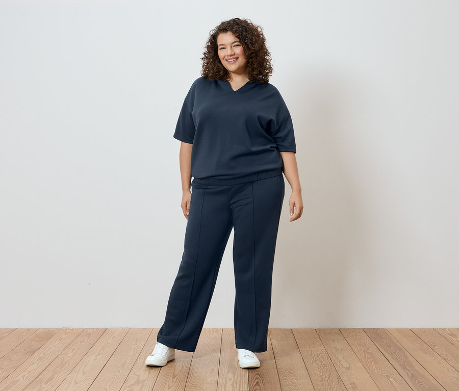 Žena s kučeravými vlasmi v tmavomodrom tričku a nohaviciach pred bielou stenou.