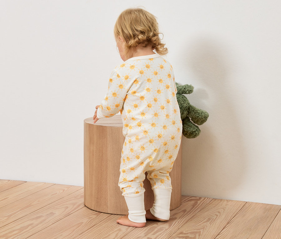 Batoľa v žlto-bielom overale so vzorom slnka stojí vedľa okrúhlej drevenej stoličky, vedľa je zelená plyšová hračka.