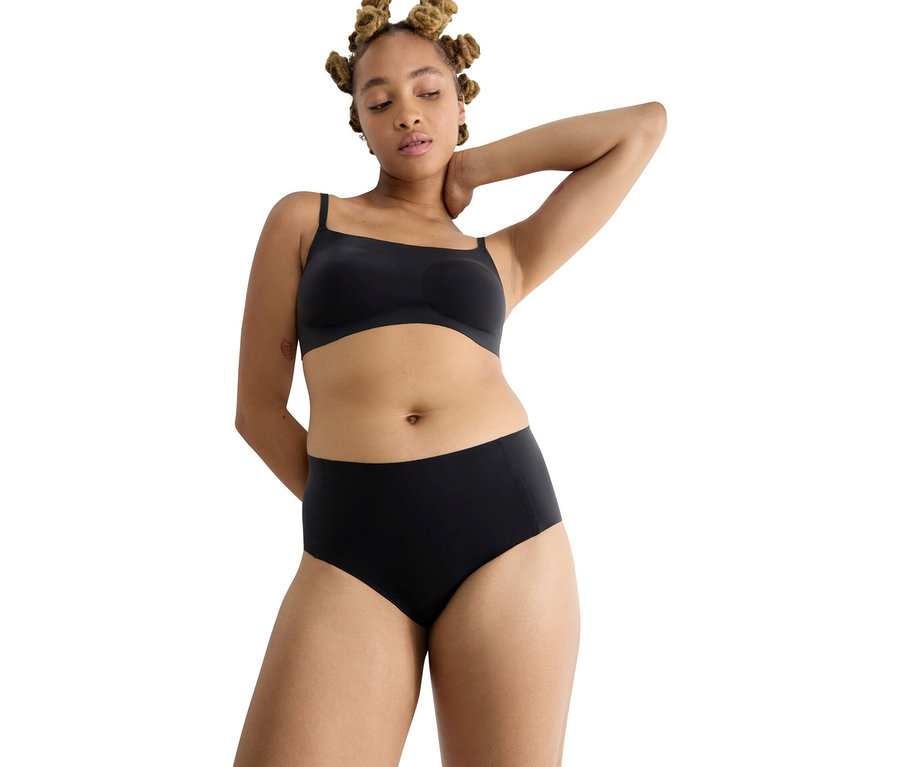 Žena s bantu uzlíkmi má na sebe čiernu podprsenku a čierne nohavičky sloggi ZERO Feel 2.0 High Waist.
