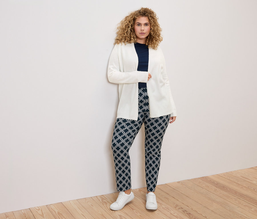 Žena s kučeravými vlasmi v bielom kardigane, tmavomodrej blúzke a vzorovaných nohaviciach stojaca pred bielou stenou.