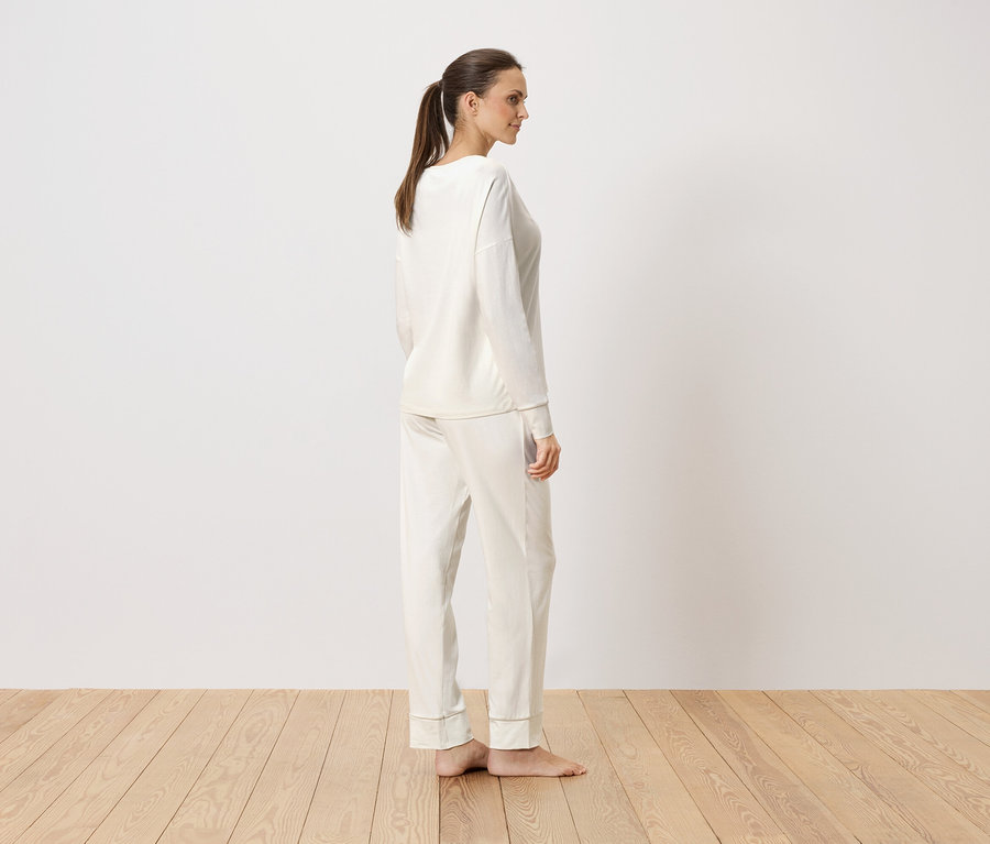 Žena stojí bokom v krémovom pyžame s dlhým rukávom pred bielou stenou.