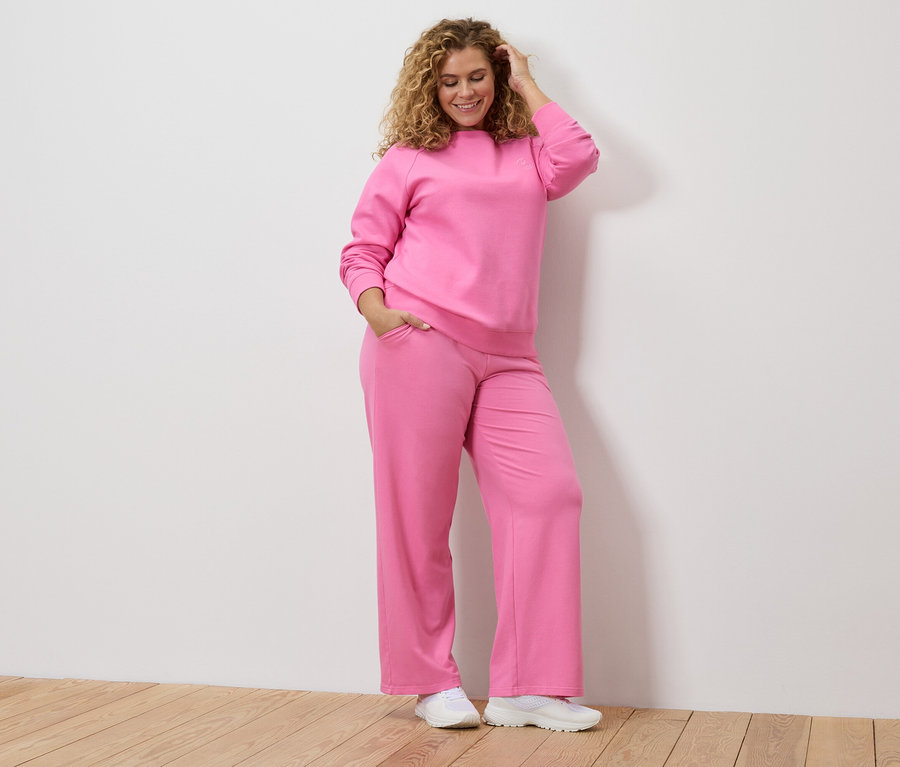 Žena s kučeravými vlasmi v ružovej teplákovej súprave a bielych teniskách si drží ruku na hlave.