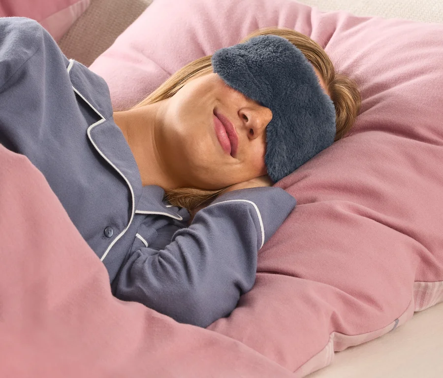 Žena s blond vlasmi pokojne spí v posteli, oblečená v modrom pyžamovom tope s bielym lemovaním a tmavosivej, huňatej maske na spanie. Je prikrytá ružovou perinou a hlavu má položenú na ružovom vankúši.