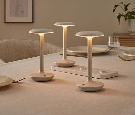 Tri minimalistické, krémové stolové lampy na prestretom jedálenskom stole, jedna z nich na nabíjacej stanici. Lampy svietia teplým svetlom.