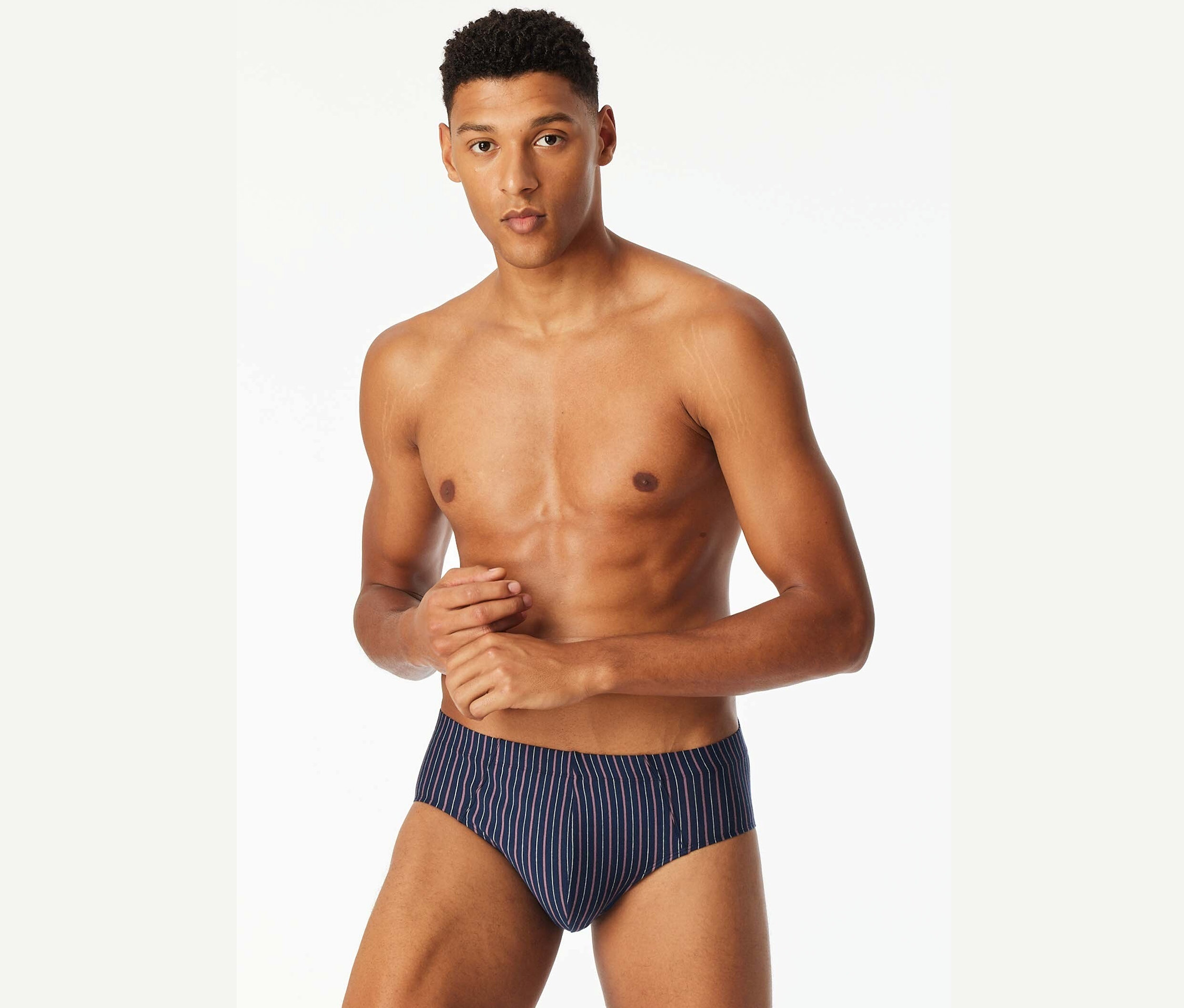Muž s odhalenou hornou časťou tela pózuje v modrých slipových plavkách s prúžkami.