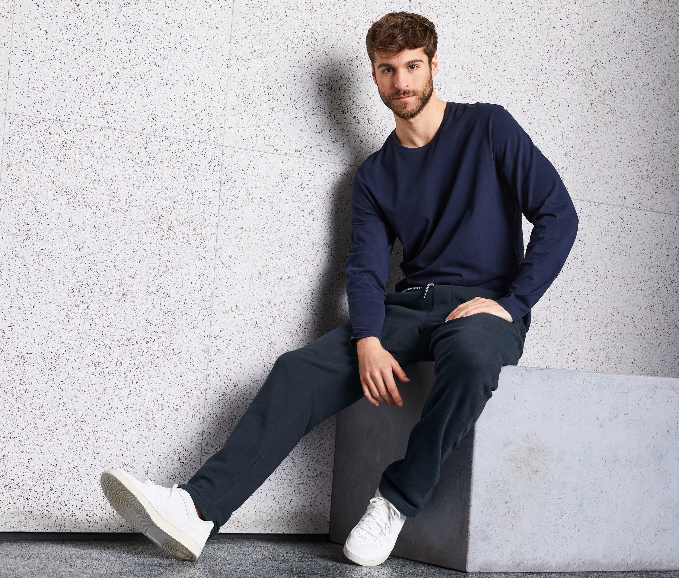 Muž sedí na betónovom bloku a má na sebe tmavomodré tričko s dlhým rukávom a teplákové nohavice chino.