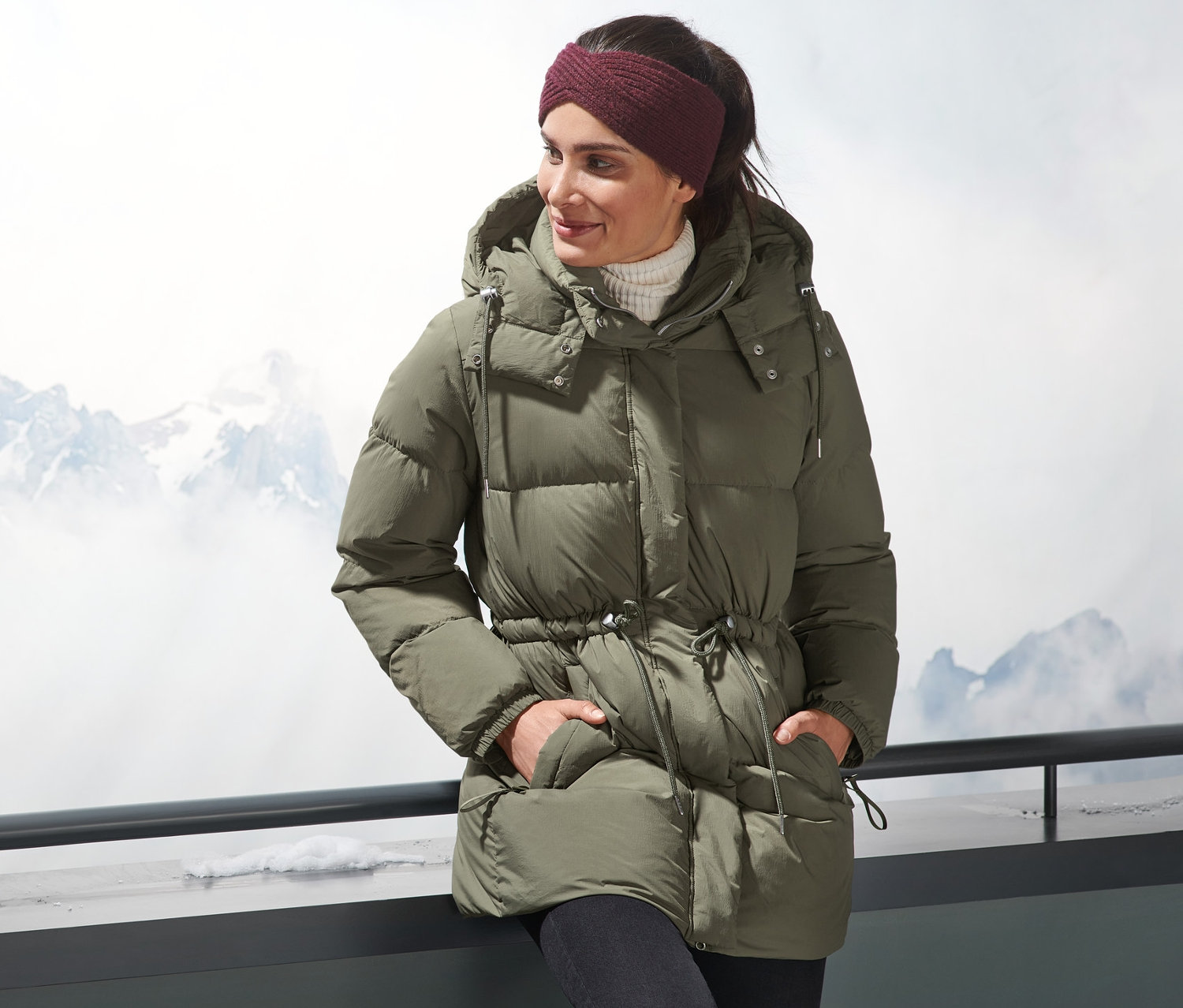 Žena v páperovej bunde stojí vonku s horami v pozadí.