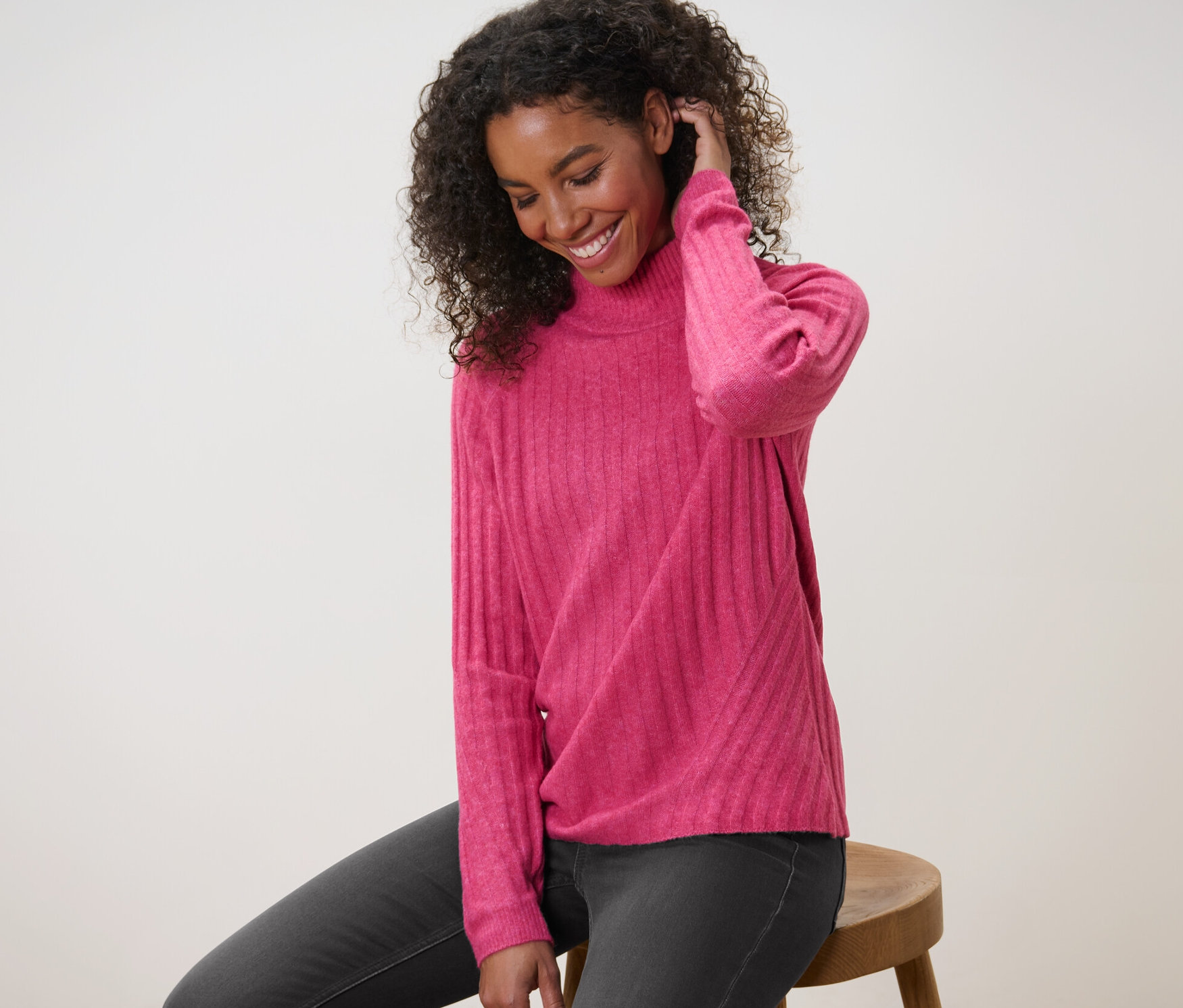 Žena sedí na stoličke a má na sebe ružový rebrovaný pletený pulóver so stojačikom.