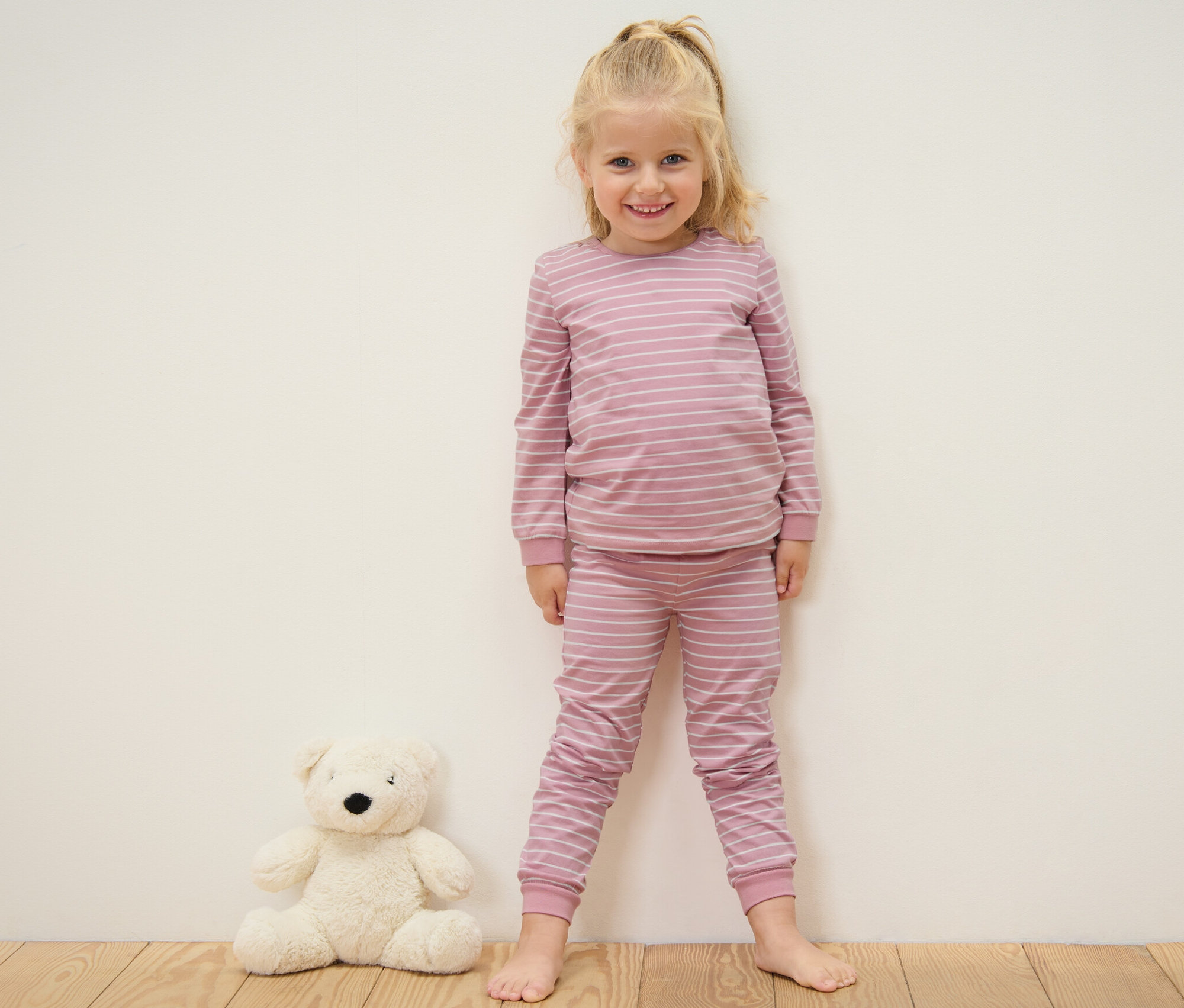Usmievajúca sa blondínka stojí pri stene v ružovom prúžkovanom pyžame a vedľa nej sedí plyšový medvedík. Detské pyžamá, 2 ks, ružové.