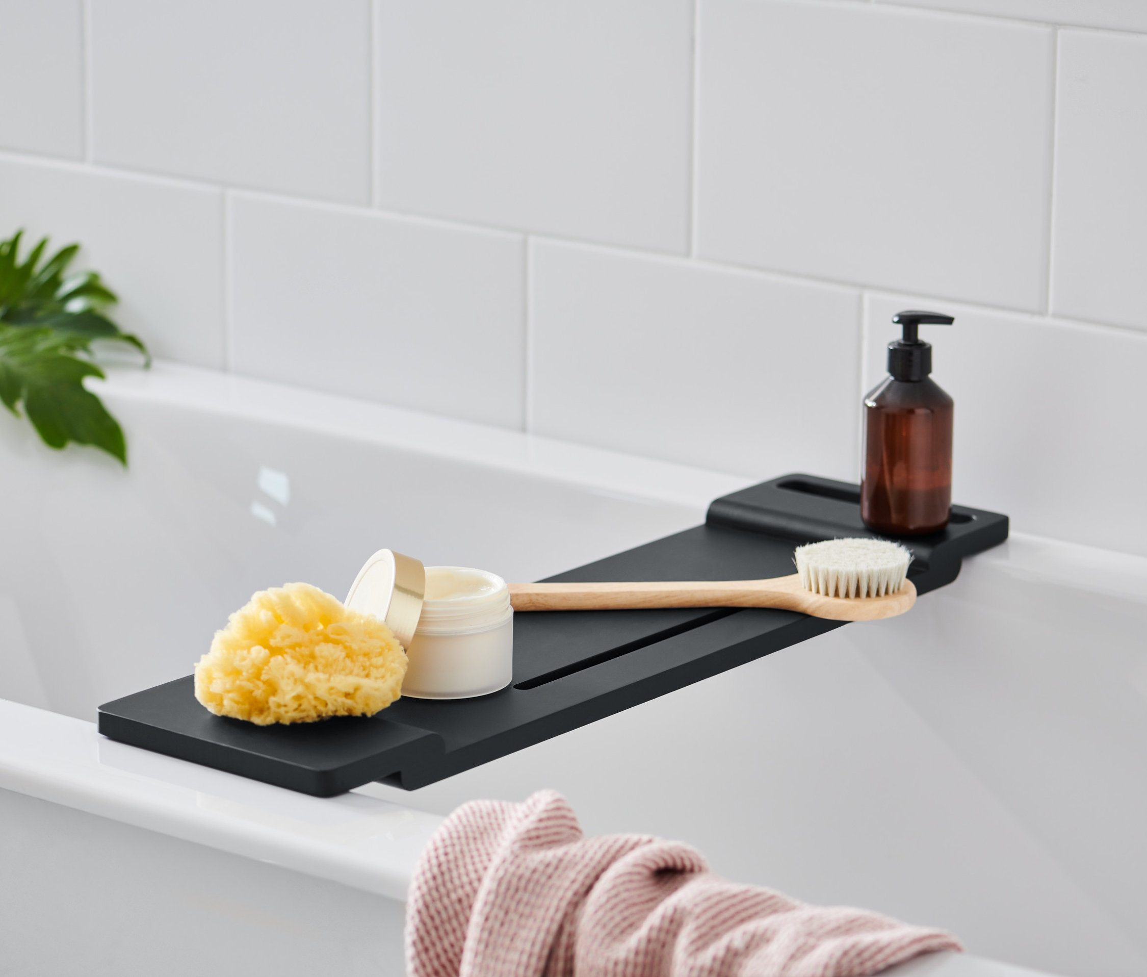 Na vani je umiestnený čierny odkladací podnos na vaňu, na ktorom leží hubka, téglik s krémom, kefa a hnedá fľaštička s pumpičkou.