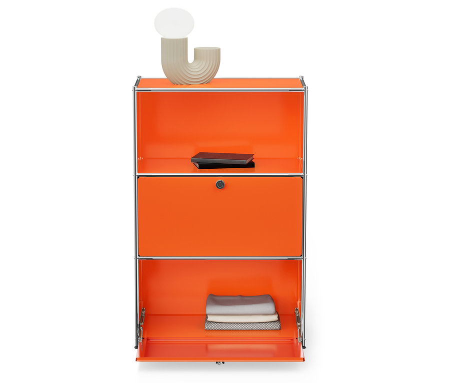 Oranžová skrinka s tromi priehradkami a strieborným rámom. Na hornej polici je svetlobéžová lampa a čierna kniha. Spodná priehradka je otvorená a sú v nej zložené látky.