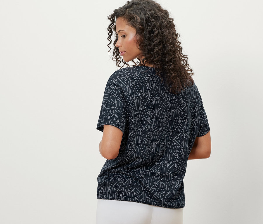 Zozadu je vidieť žena s kučeravými vlasmi, ktorá má na sebe tmavomodré spadnuté blúzkové tričko a biele nohavice.