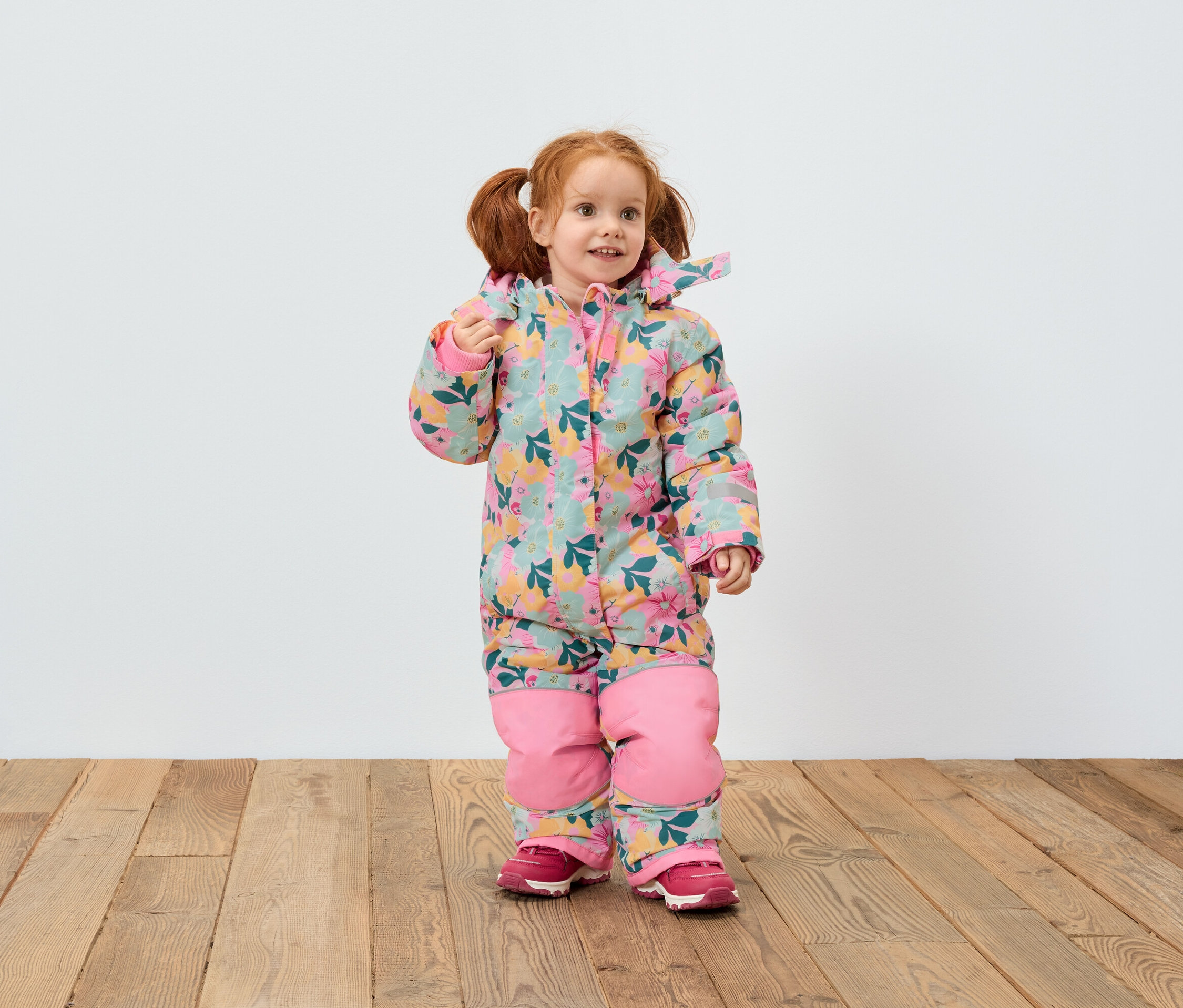 Dievča s ryšavými vlasmi v kvetinovom overale a ružových topánkach stojí na drevenej podlahe pred bielou stenou.