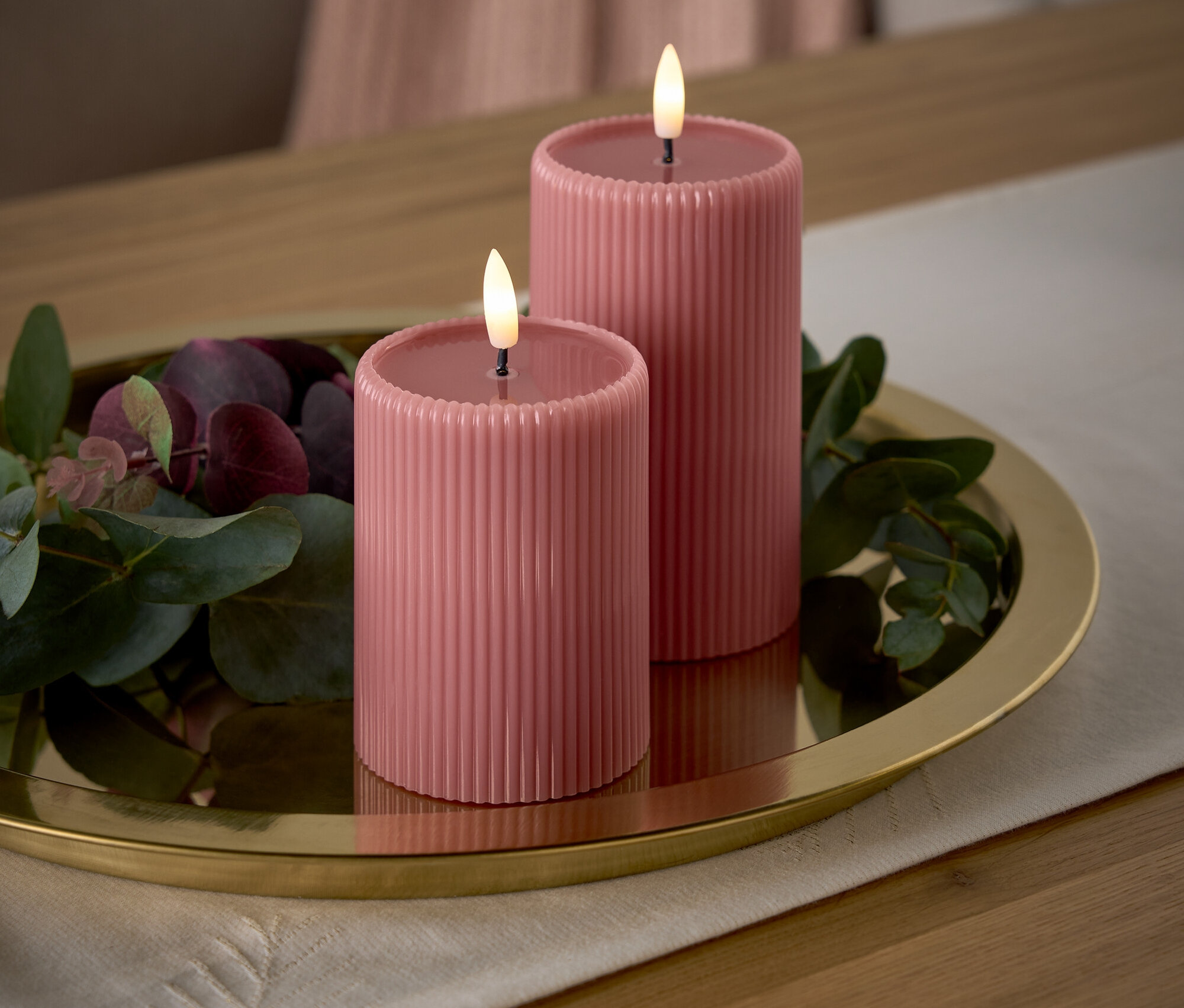 Dve ružové sviečky s plameňmi na zlatom podnose s dekoratívnymi vetvičkami na stole.