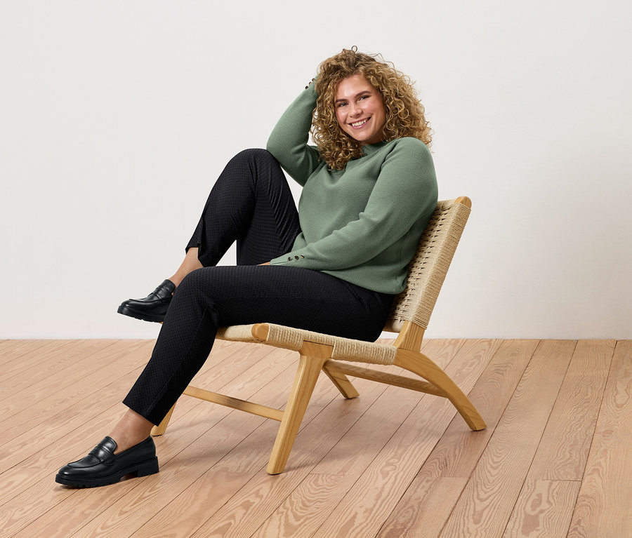 Žena s kučeravými vlasmi sedí na ratanovom kresle, má na sebe zelený sveter, čierne nohavice a topánky.