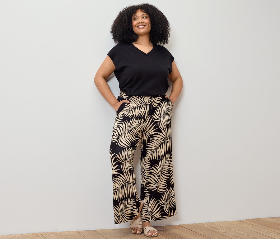 Žena pózuje v čiernom pulóvri z jemného úpletu s krátkymi rukávmi a nohaviciach v štýle palazzo s potlačou.