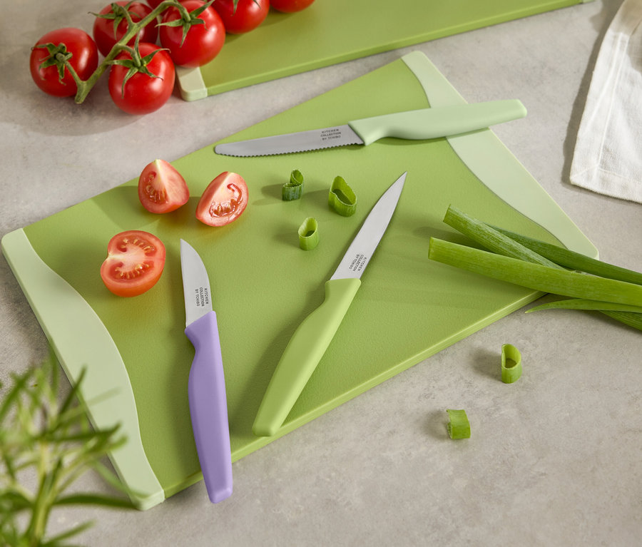 Zelená doska na krájanie s nakrájanými paradajkami, jarnou cibuľkou a tromi nožmi s farebnými rukoväťami.