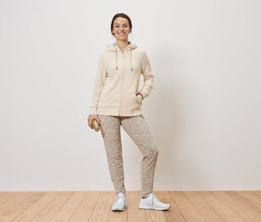 Žena stojí a drží zlatú fľašu. Má na sebe béžovú mikinu na zips, vzorované nohavice a biele tenisky.