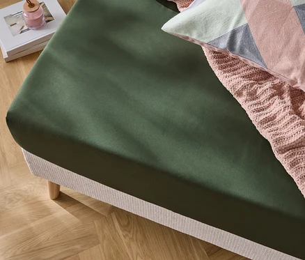 Detailný záber na posteľ s olivovo zelenou plachtou, ružovou pletenou dekou a ozdobným vankúšom.