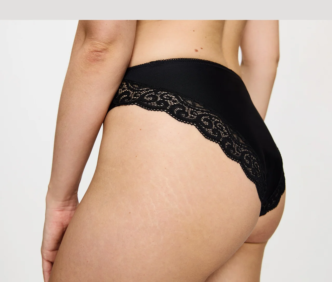Detailný záber zozadu na osobu, ktorá má na sebe čierne nohavičky Triumph Amourette Tai s čipkovaným lemom.