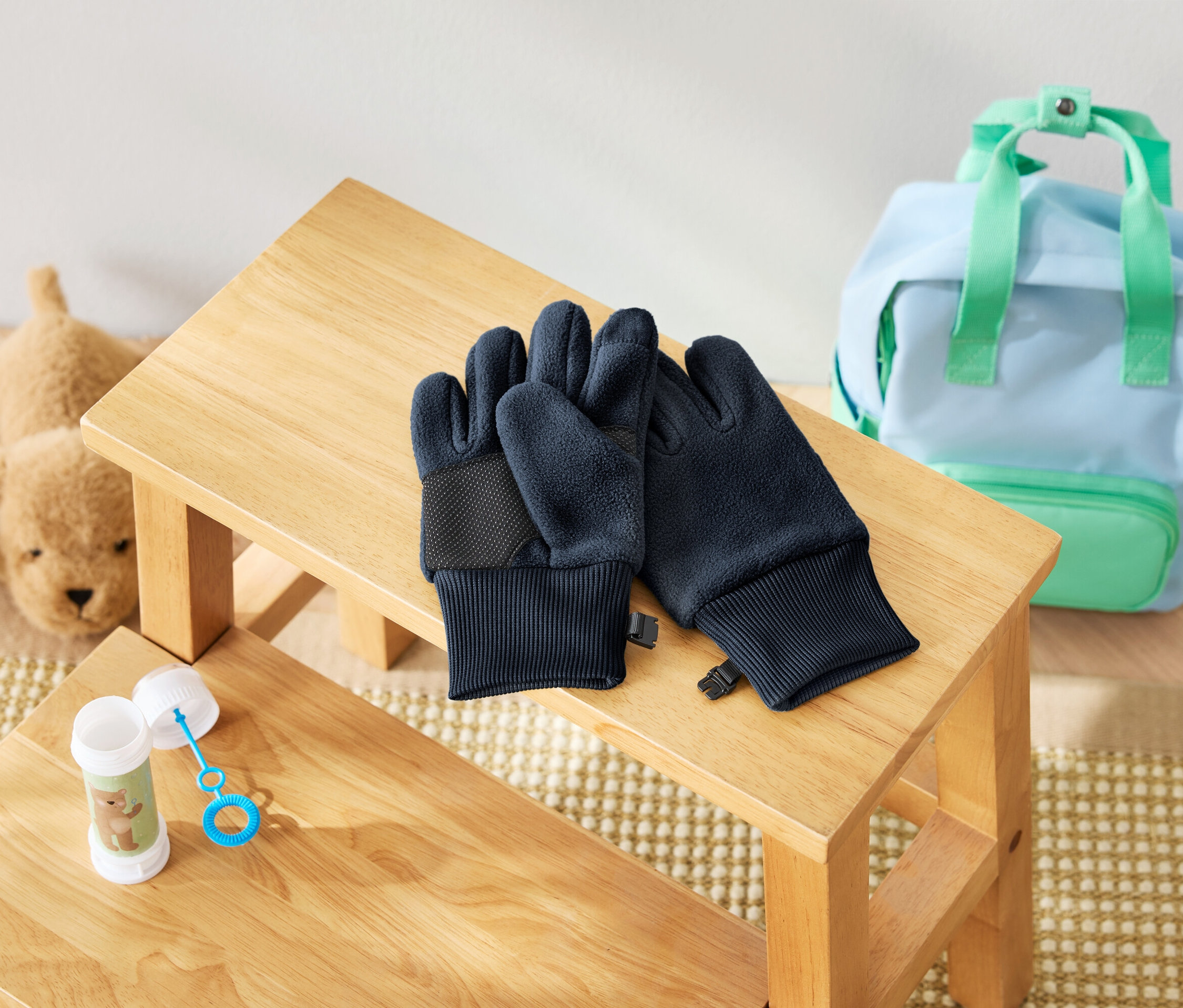 Na drevenej stoličke leží pár tmavomodrých rukavíc, vedľa nich je fľaša s bublifukom a plyšový pes. V pozadí je svetlomodrý batoh s mätovo zelenými detailmi.