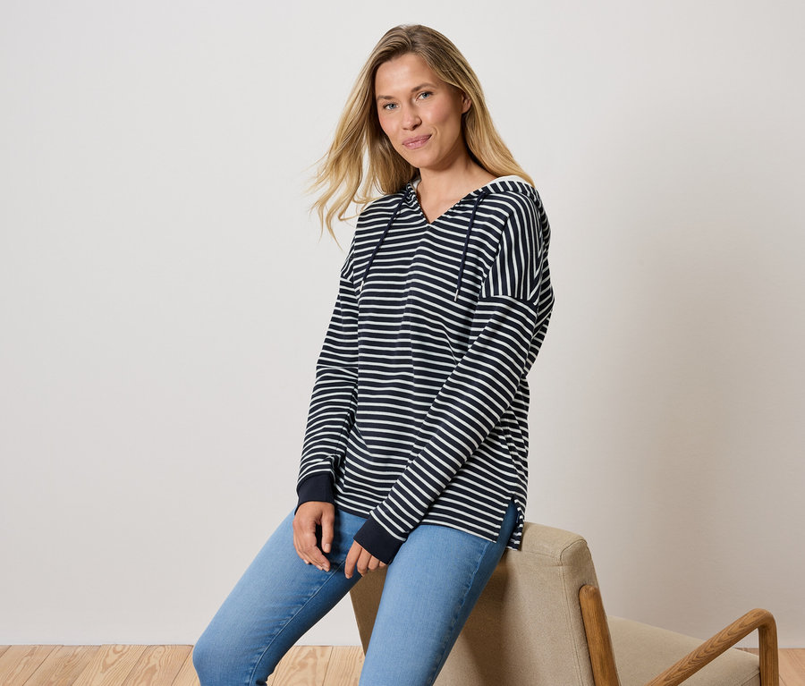 Žena sedí na stoličke, oblečená v modrých džínsoch a pruhovanej mikine s kapucňou.