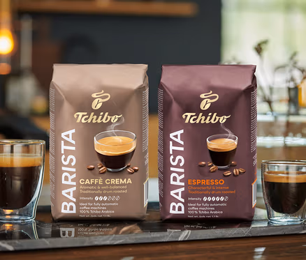 Dva poháre kávy a dve balenia zrnkovej kávy Tchibo BARISTA Caffè Crema stoja na mramorovej doske.