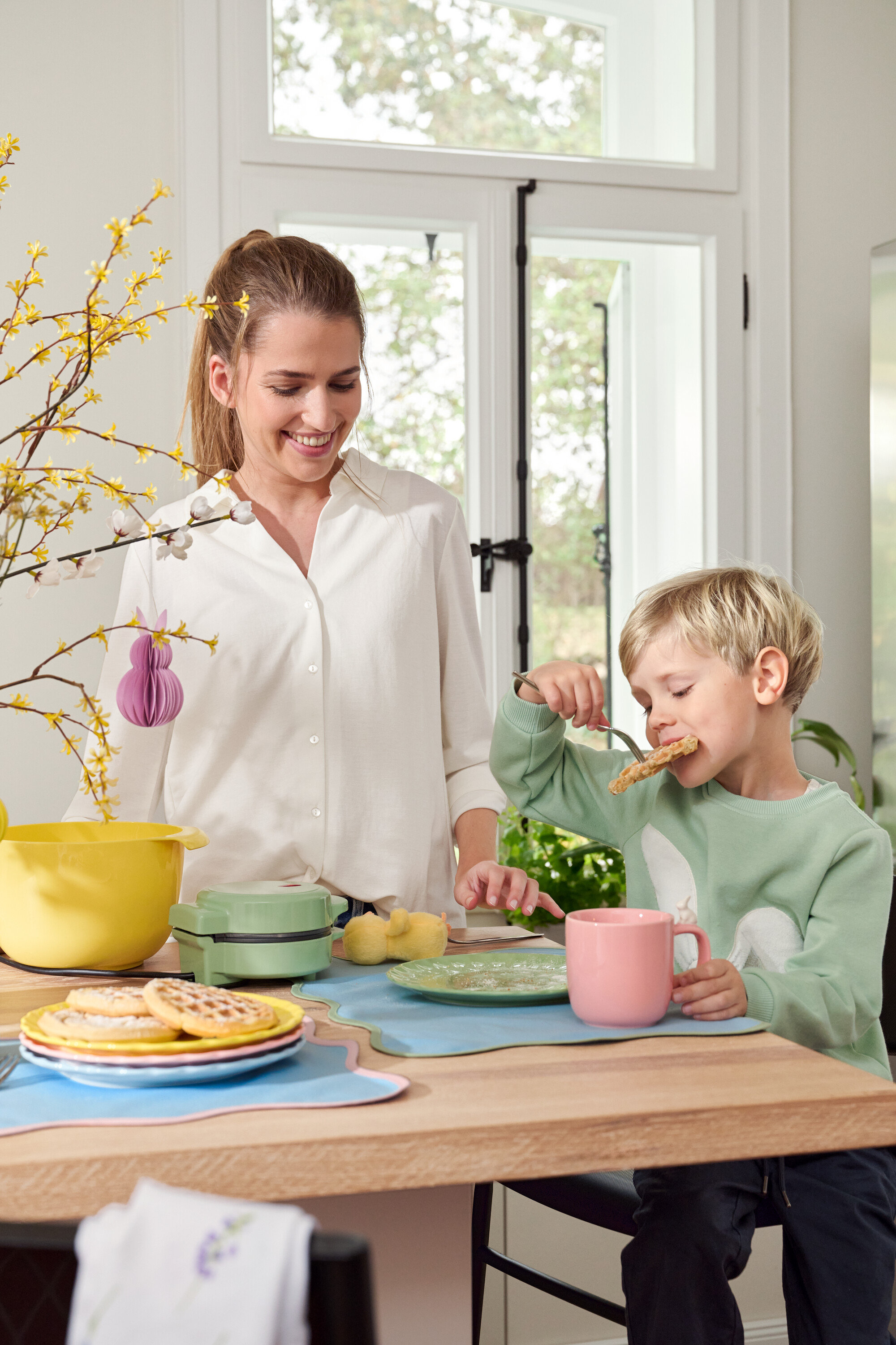 Žena a dieťa pri stole. Dieťa je vafle vidličkou, žena sa usmieva.