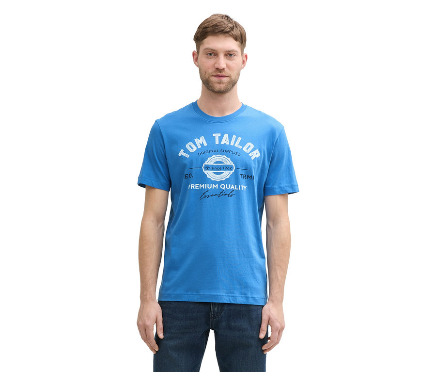 Muž má na sebe tričko s logom Tom Tailor, sapphire blue a džínsy.