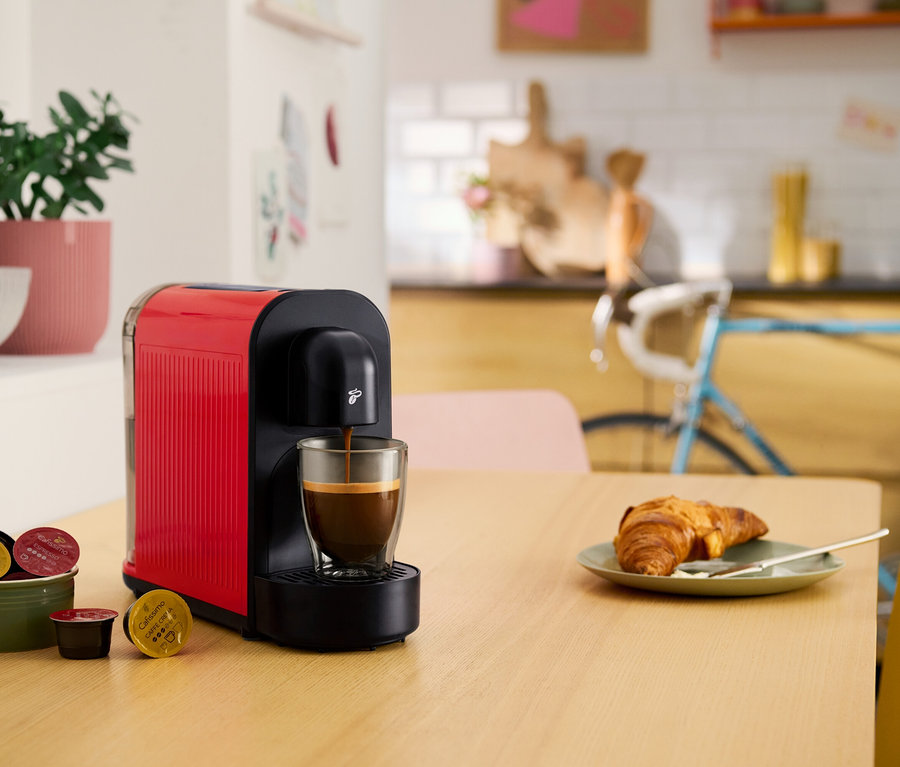 Červený kapsulový kávovar plní šálku kávou. Na stole je croissant, miska s kávovými kapsulami a črepníková rastlina.