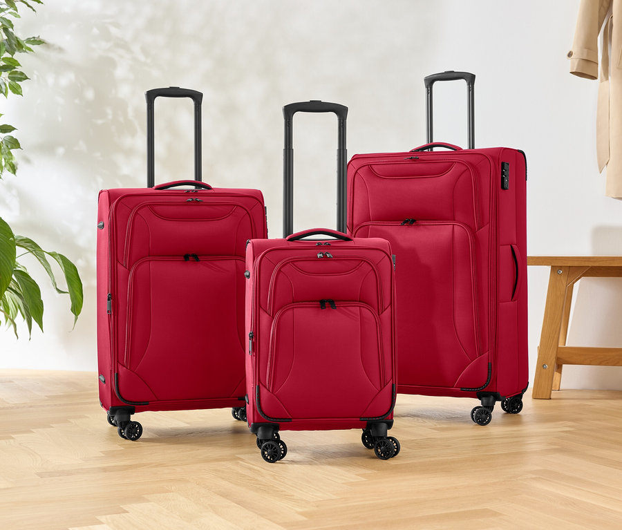 Tri červené kufre rôznych veľkostí stoja na drevenej podlahe, vedľa rastliny a lavice.
