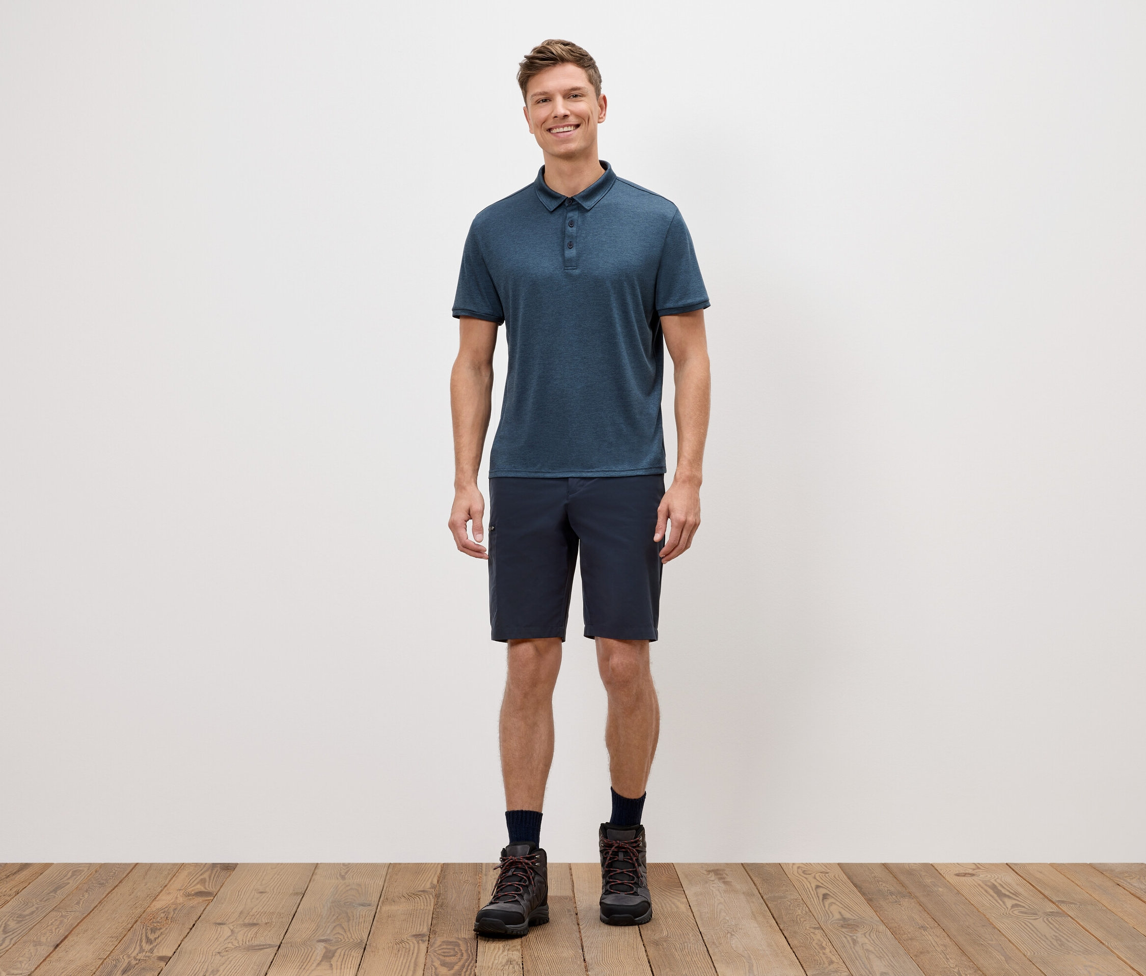 Muž stojí pred bielou stenou a má na sebe modré polokošeľu, krátke nohavice a trekové topánky.