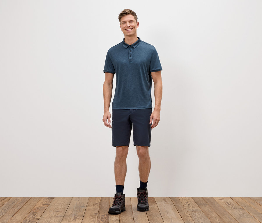 Muž stojí pred bielou stenou a má na sebe modré polokošeľu, krátke nohavice a trekové topánky.