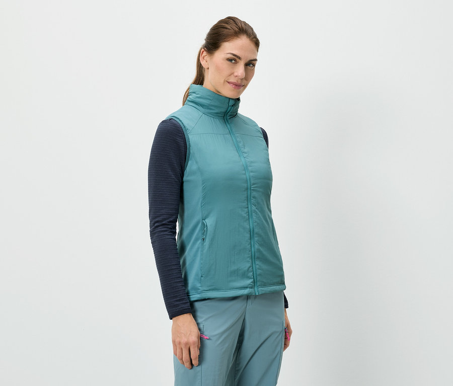 Žena stojí a má na sebe modrú vestu cez košeľu s dlhými rukávmi, v kombinácii s modrými nohavicami.