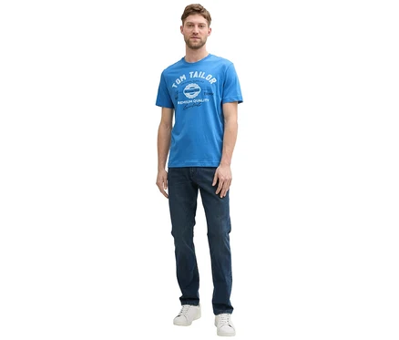 Muž v džínsoch a bielych teniskách má na sebe tričko s logom Tom Tailor vo farbe sapphire blue.