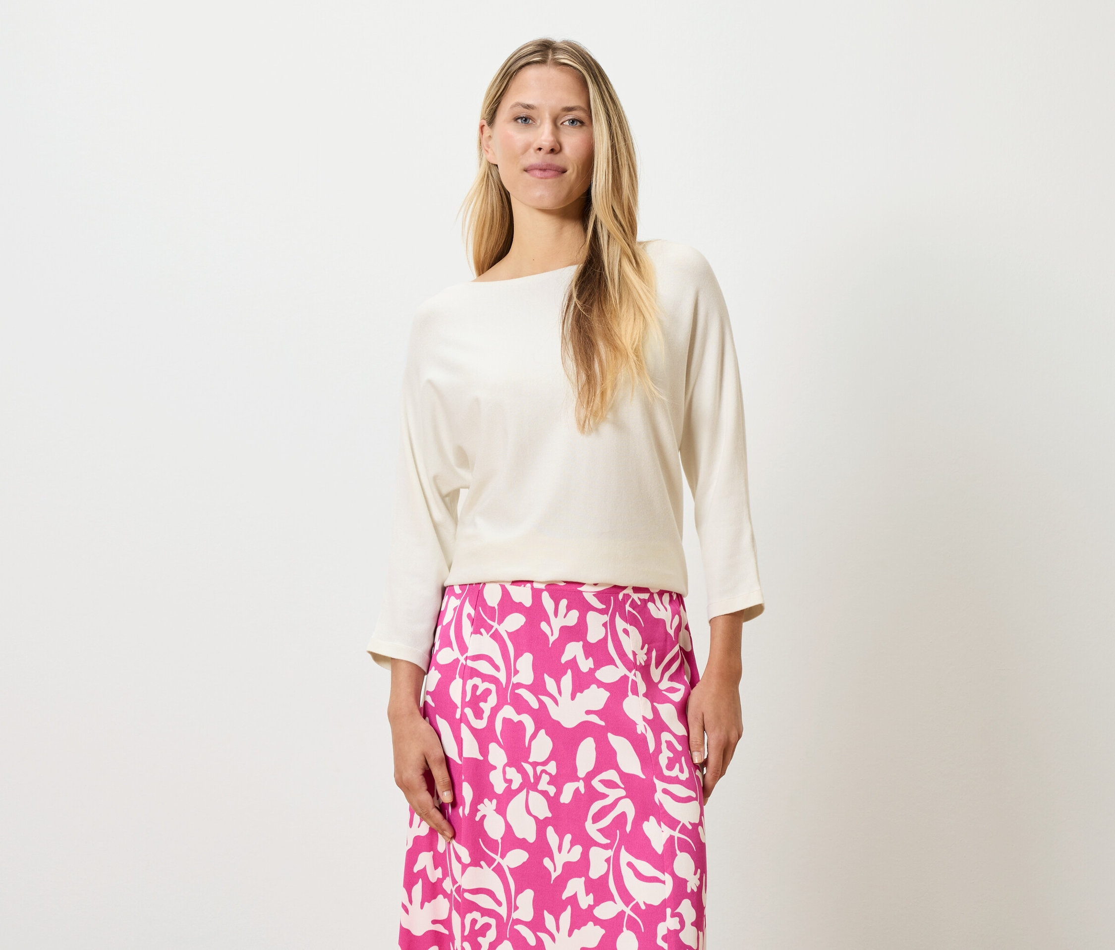 Žena s blond vlasmi má na sebe krémový top a ružovú sukňu s kvetinovou potlačou.