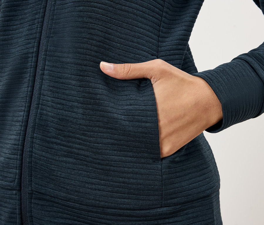 Detail osoby v tmavomodrej štruktúrovanej bunde, ktorá si dáva ruku do vrecka bundy.