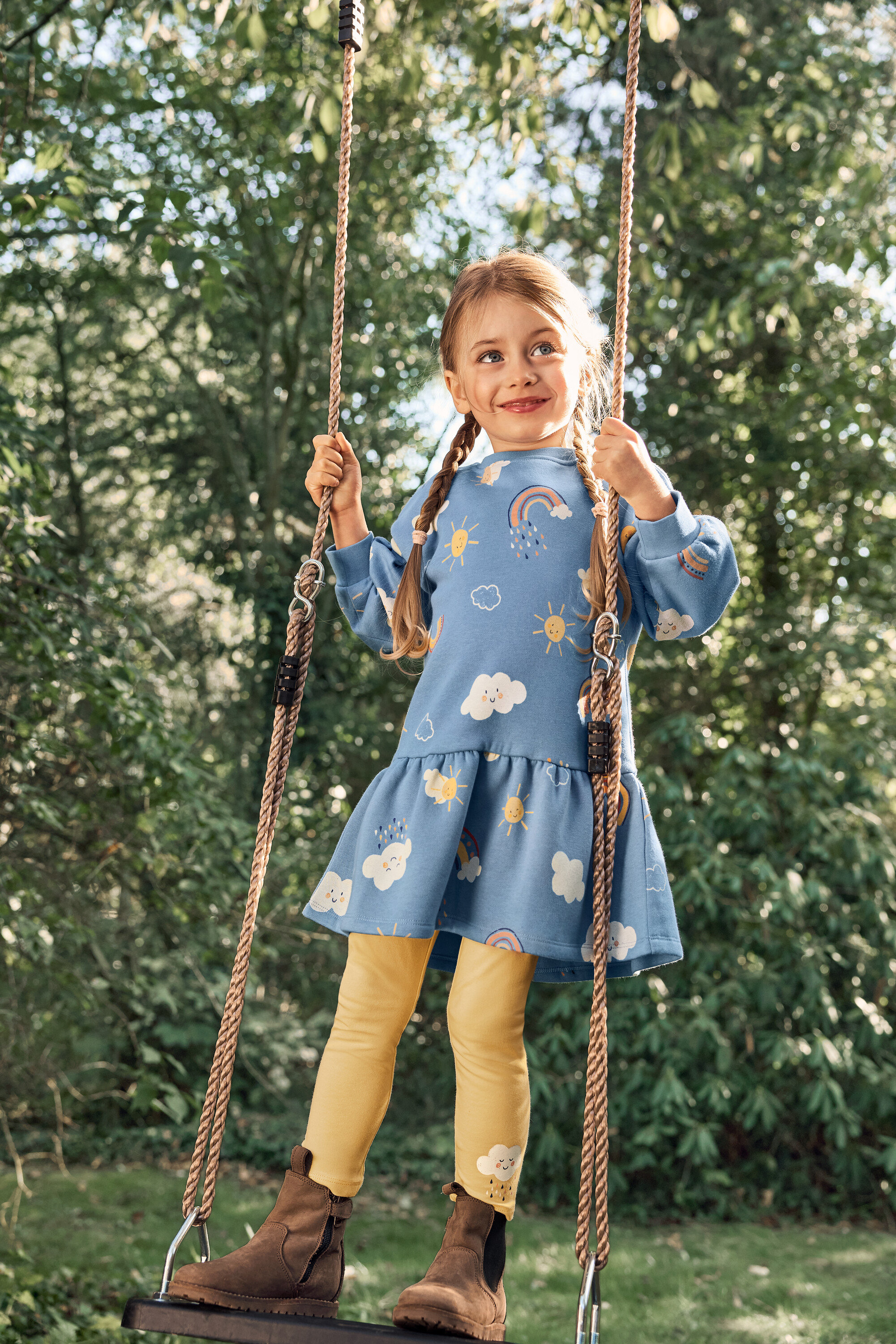 Dievča so zapletenými vlasmi sa húpe na hojdačke, má na sebe modré šaty so vzormi slnka, oblakov a dúhy, žlté legíny a hnedé topánky.