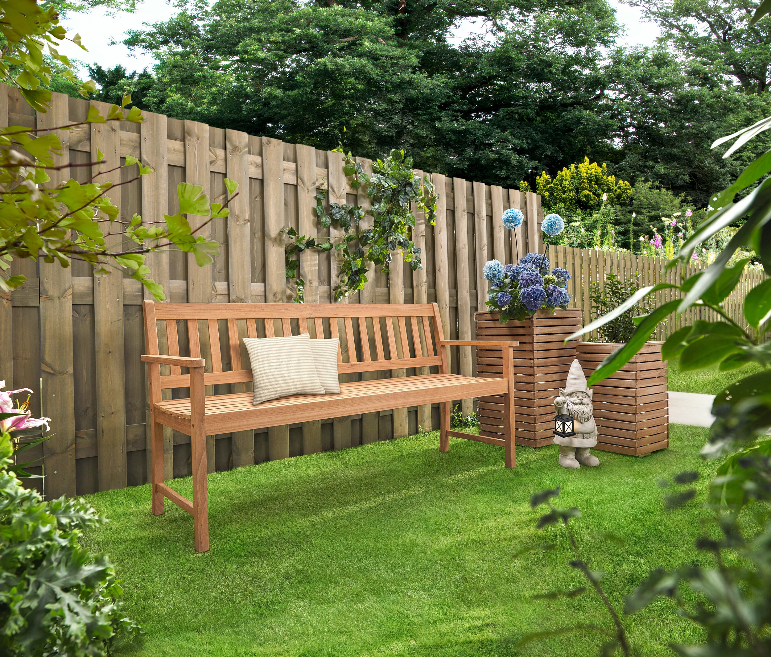 Drevená lavica s vankúšmi stojí na trávniku. Za ňou stoja dva stĺpy na kvety »Tinus« s modrými kvetmi.